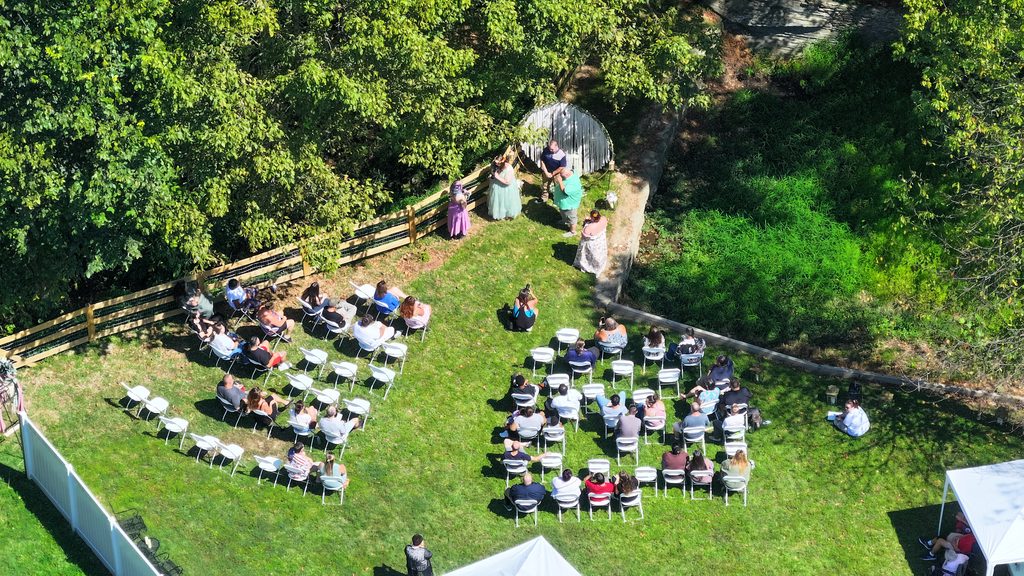 Top-down aerial view of a backyard wedding ceremony: guests in rows of white chairs on the lawn, the couple and officiant under a fringed arch by a wooden fence.