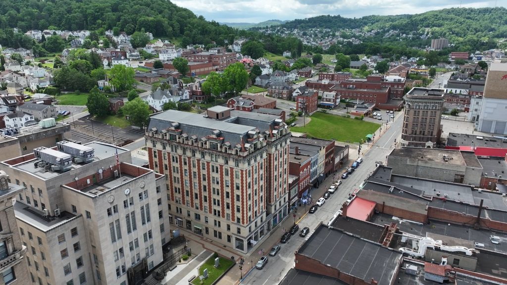 Aerial overview of a small-city downtown: a historic brick-and-stone high-rise, flat commercial roofs, and a main street, with wooded hills and neighborhoods behind.