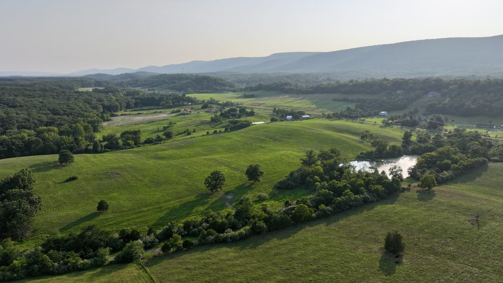 Golden-hour aerial view over a green valley: rolling pasture, tree lines, a small pond, and a hazy mountain ridge on the horizon.
