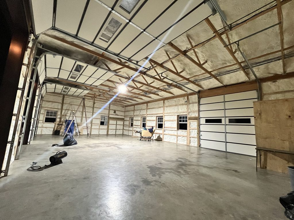 Empty pole barn interior after spray foam: exposed trusses, roll-up doors, a light flaring across the ceiling.