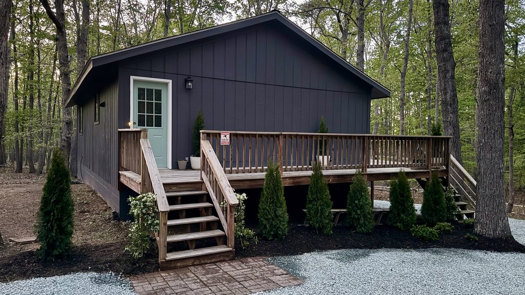 Same cabin after renovation: dark charcoal siding, mint door, new lighting, stained deck, fresh mulch beds with arborvitae, gravel and a brick paver walk.