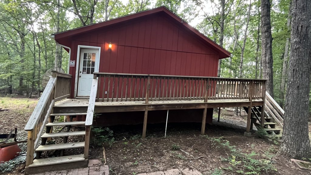 Cabin before renovation: red board-and-batten siding, weathered deck and stairs, bare dirt yard in the woods.