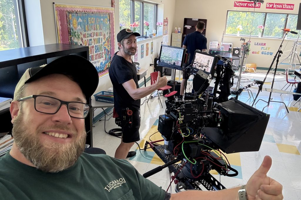 Joey Walker giving a thumbs up beside a fully built cinema camera rig with monitors on a classroom set, crew member and grip gear behind.