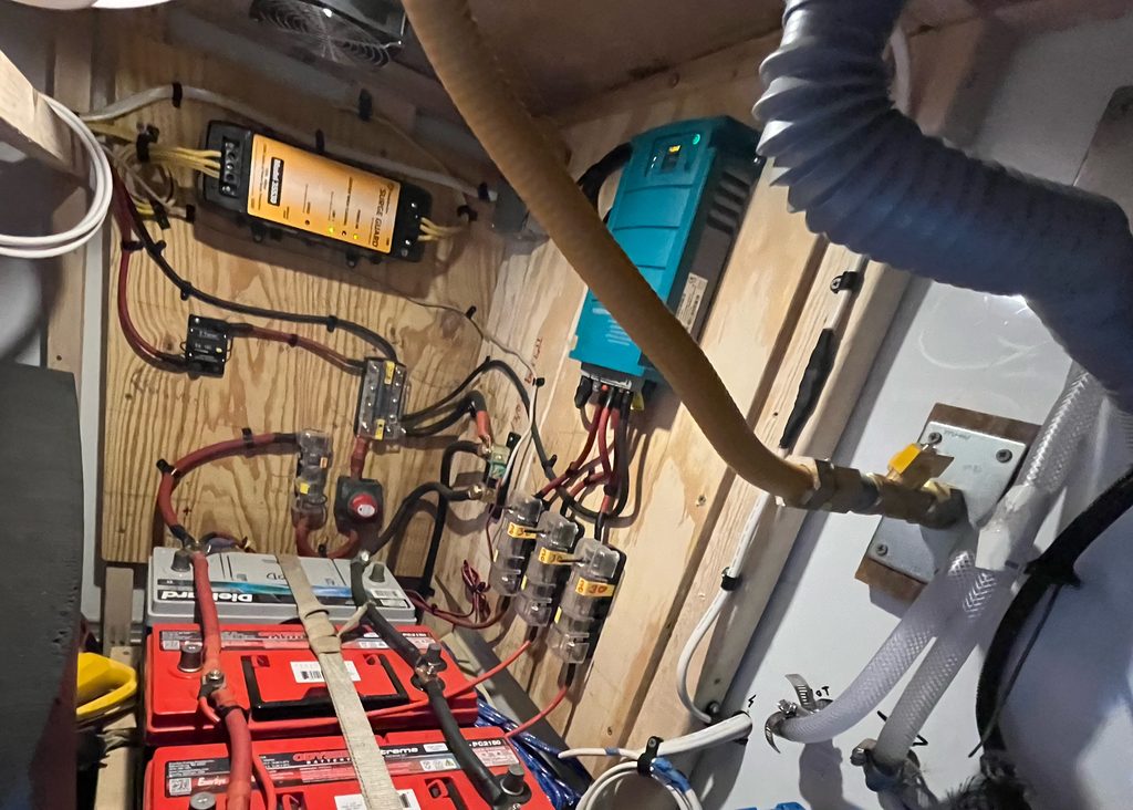 Custom off-grid electrical bay: two red deep-cycle batteries, bus bars, fuses, a blue charger/inverter, and neat wiring on plywood.