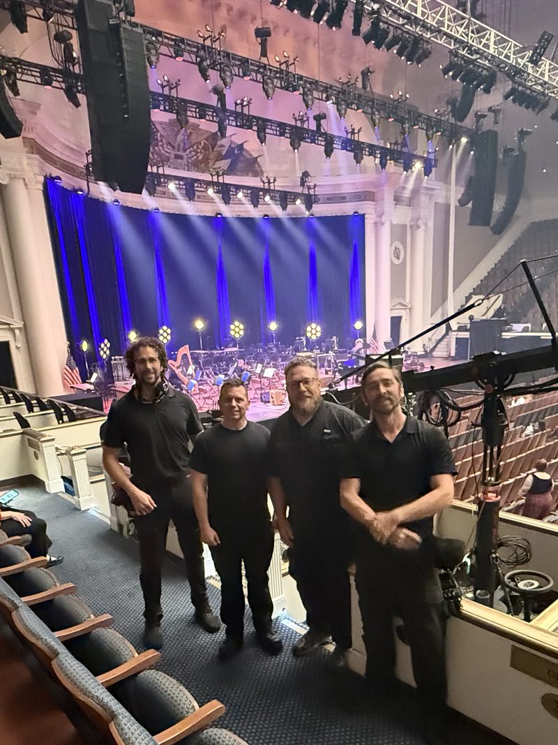 Four-person crew in black — Joey Walker third from left — in a concert-hall balcony box with the stage lighting rig, orchestra setup, and a camera crane arm.