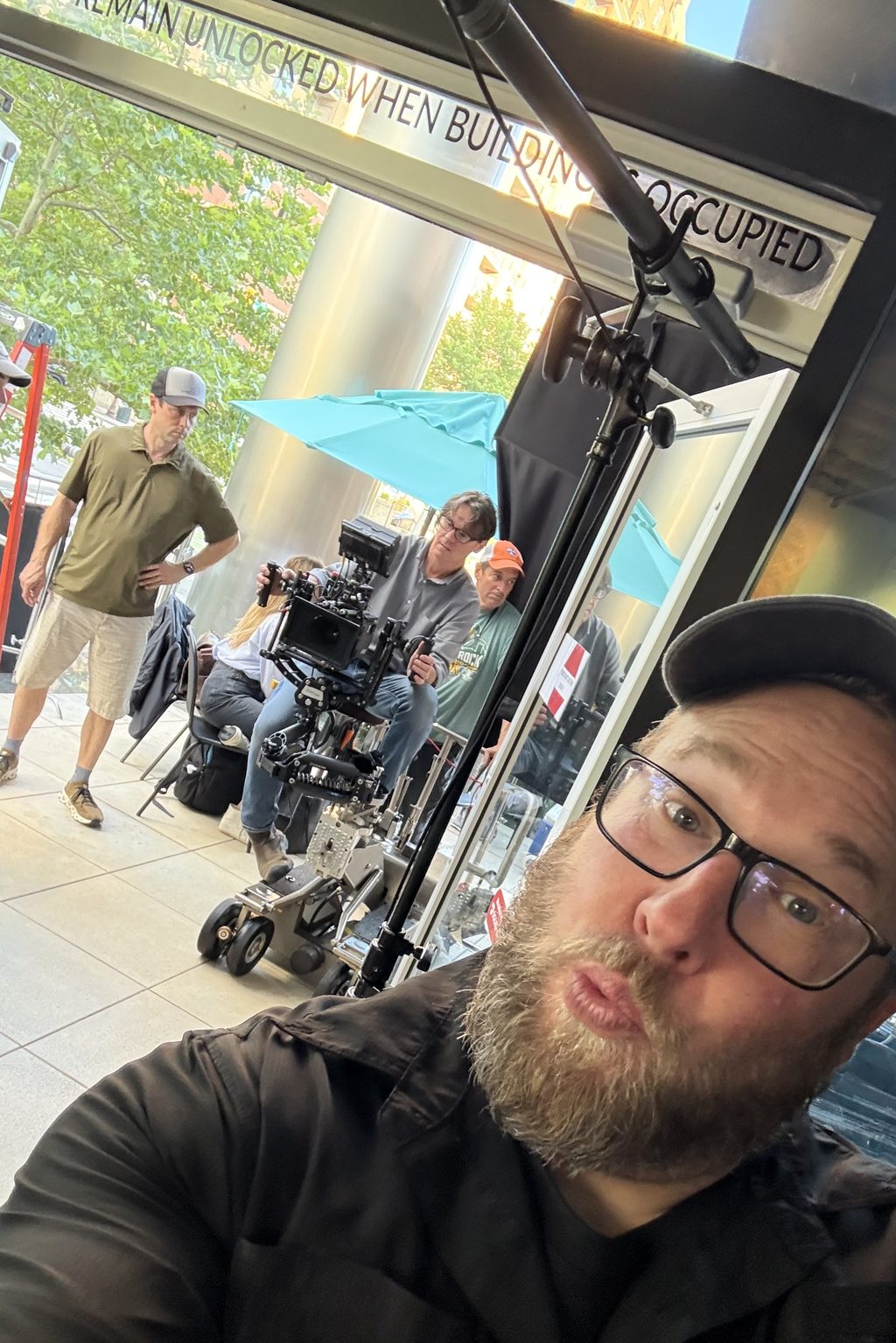 Joey Walker in the foreground on a city sidewalk; behind him a camera operator rides a dolly with a cinema camera, crew and a boom pole nearby.