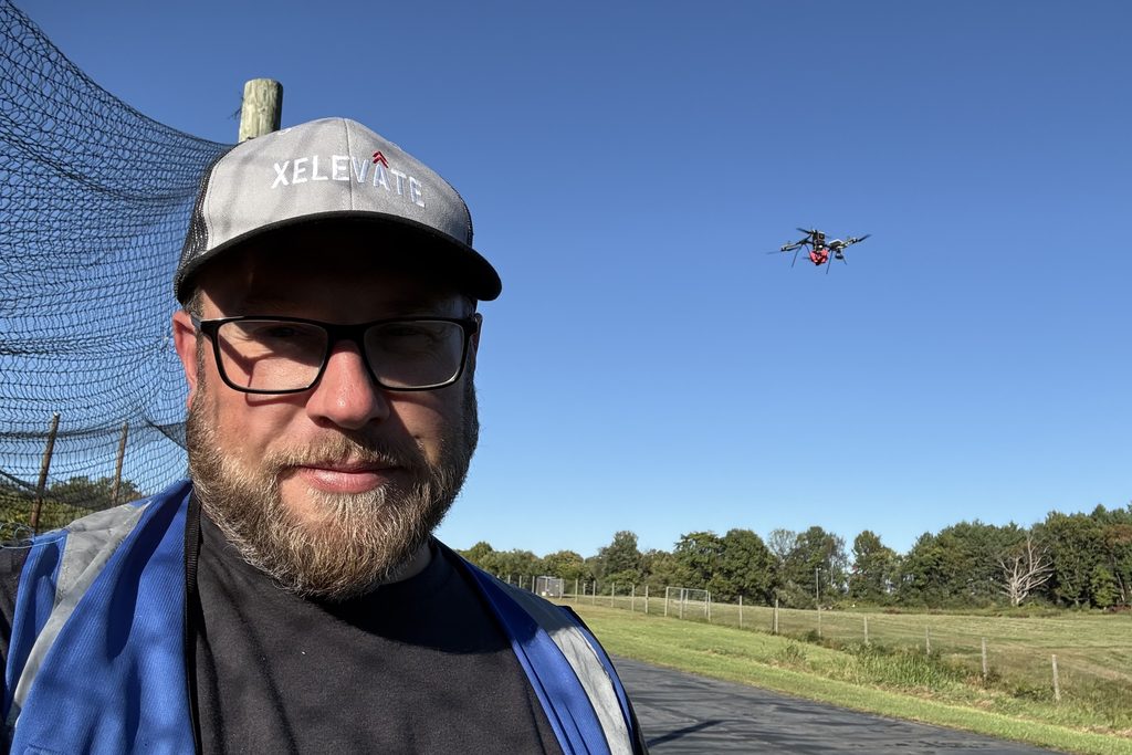 Joey Walker in a hi-vis vest with a drone flying in clear blue sky over a field behind him.