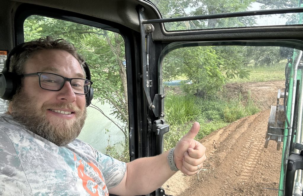 Joey Walker in ear protection giving a thumbs up from an excavator cab, a freshly cut dirt path and the bucket visible through the door.