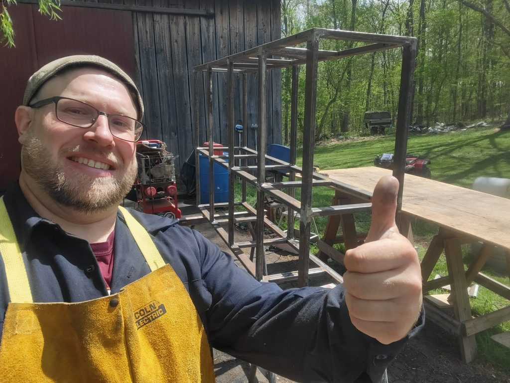 Joey Walker in a leather welding apron giving a thumbs up next to the bare welded steel tube frame of the front box.