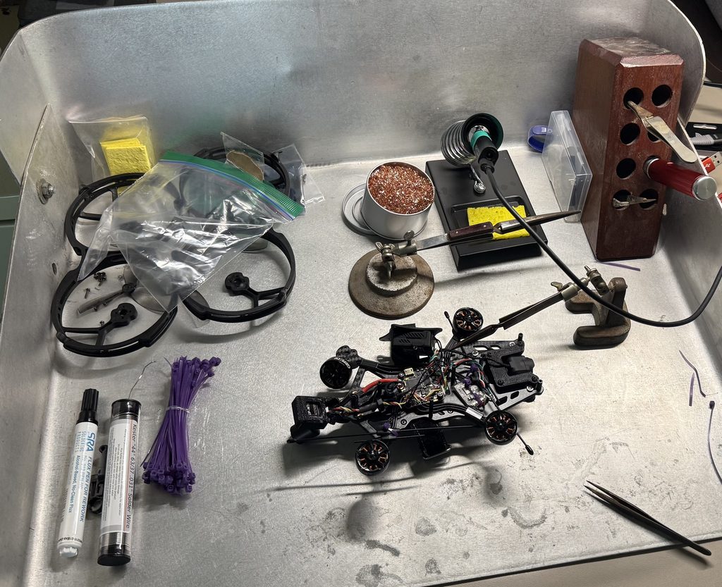 An FPV drone torn down on an aluminum tray beside a soldering station, tweezers, solder, zip ties, and spare prop guards.