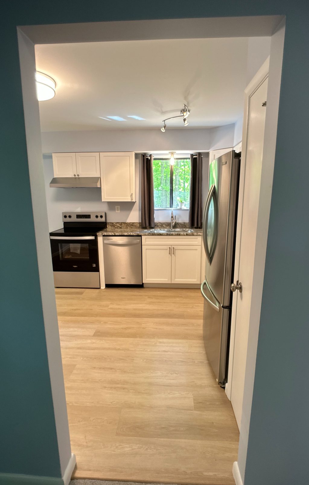 Finished kitchen seen through a doorway: white shaker cabinets, range and hood, dishwasher, granite counter with the sink under the window, a stainless French-door fridge, and LVP floor.
