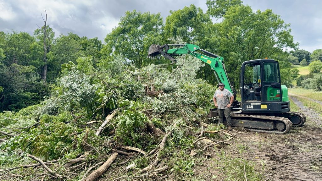 Joey Walker standing beside an E45 mini excavator next to a large pile of cleared brush and trees, a dirt track to the right.