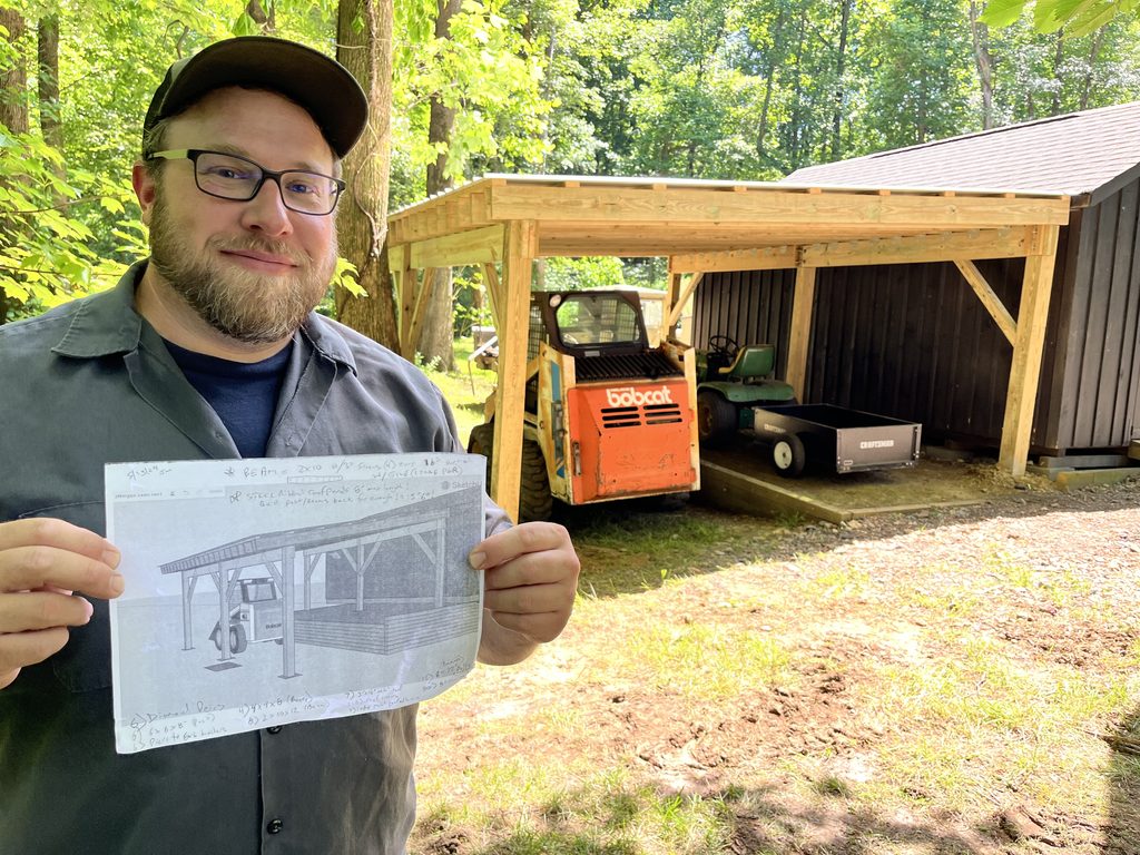 Joey Walker smiling and holding a printed SketchUp drawing of the lean-to in front of the finished timber lean-to, an orange Bobcat skid steer and a mower parked under it.