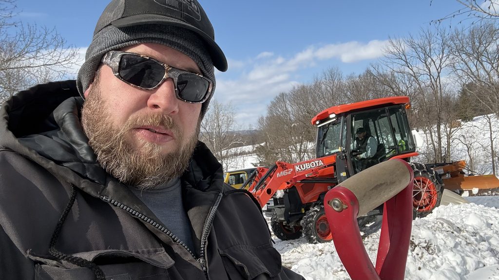 Joey Walker in a beanie and sunglasses holding a red shovel, an orange Kubota M62 loader with a plow blade behind him in deep snow.