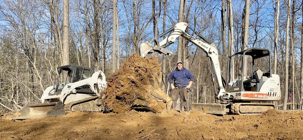 Joey Walker standing between a white Bobcat skid steer and a Bobcat 329 mini excavator holding a giant root-ball stump, on a cleared winter forest site under blue sky.