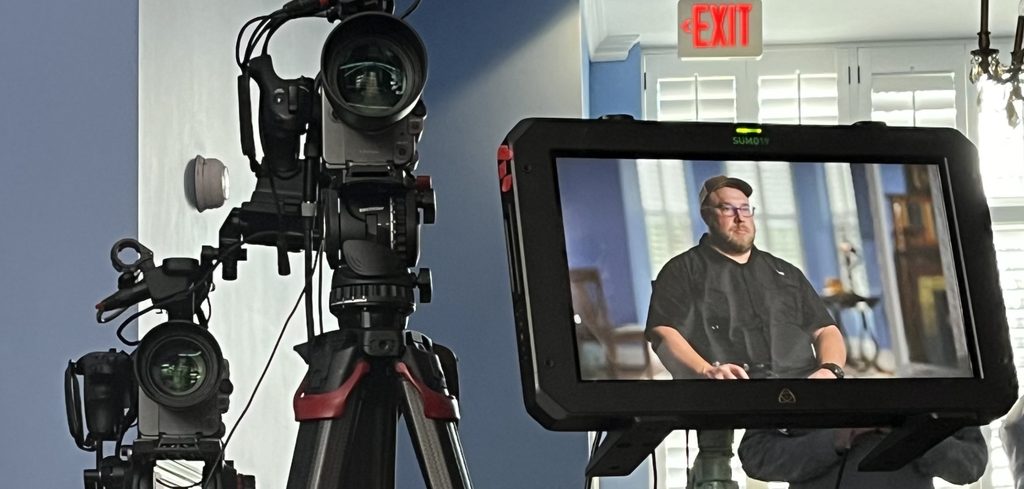 Two broadcast cameras on tripods beside an Atomos Sumo 19 monitor showing Joey Walker seated for an interview, in a bright room with shutters.